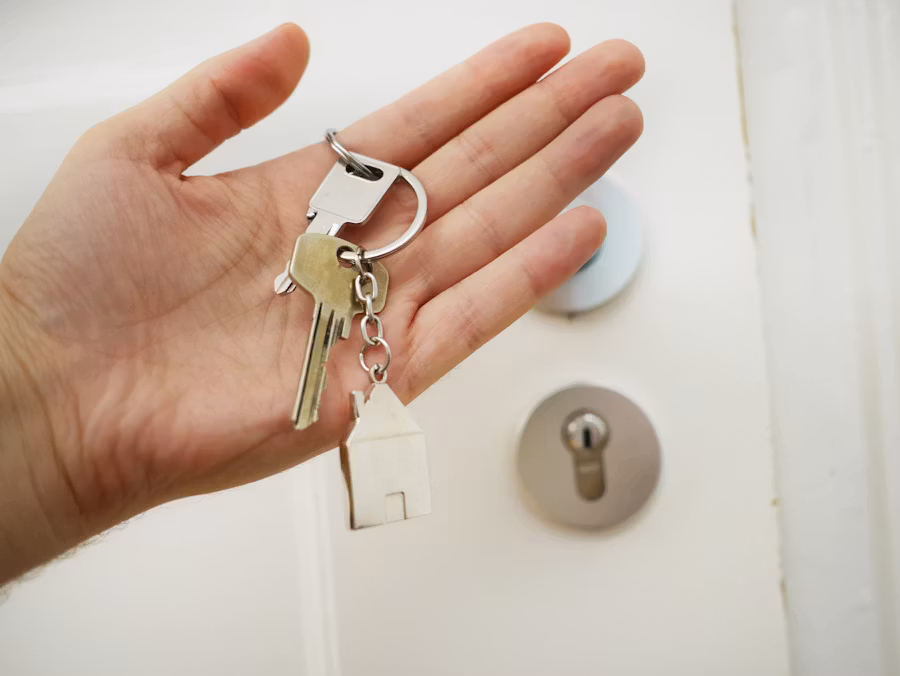 An open hand holding a key ring with a small house charm in front of a white flat door
